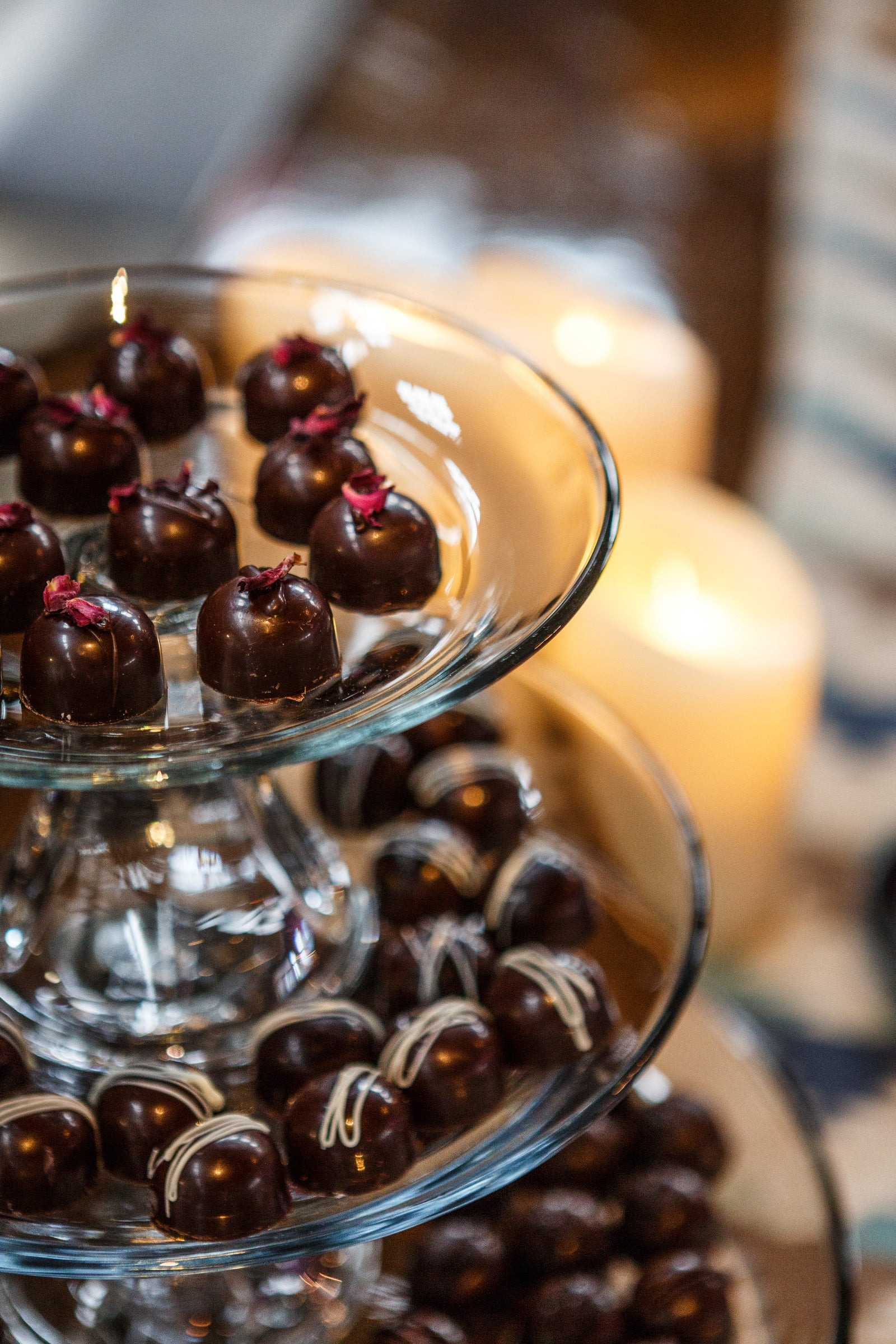 A tiered glass stand holds assorted chocolate truffles, some topped with dried flowers, with softly glowing candles blurred in the background.
