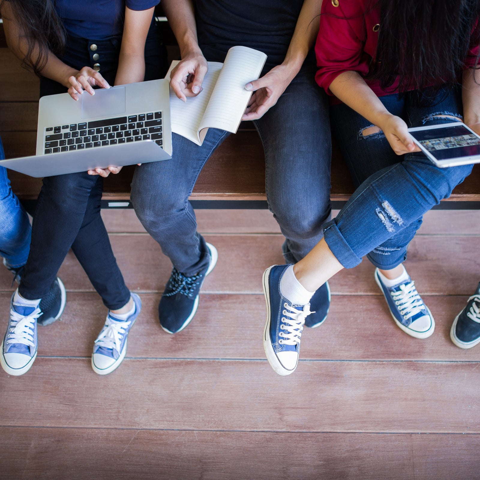 Three people sit on a bench with a laptop, notebook, and tablet, working together. Only their lower bodies and hands are visible, showing casual clothing and sneakers.