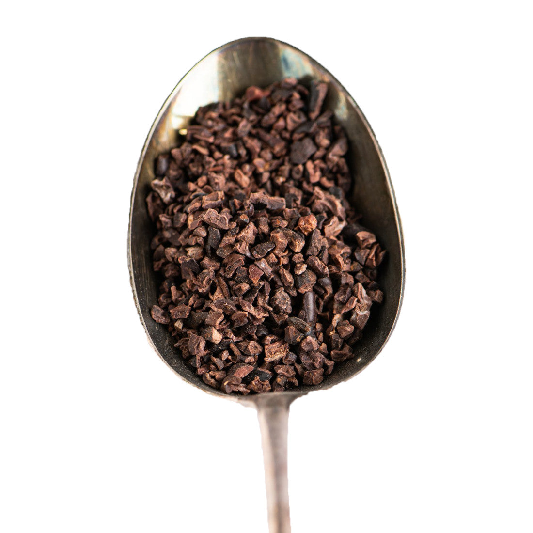A close-up of a spoon filled with small, dark brown cacao nibs, isolated on a white background.