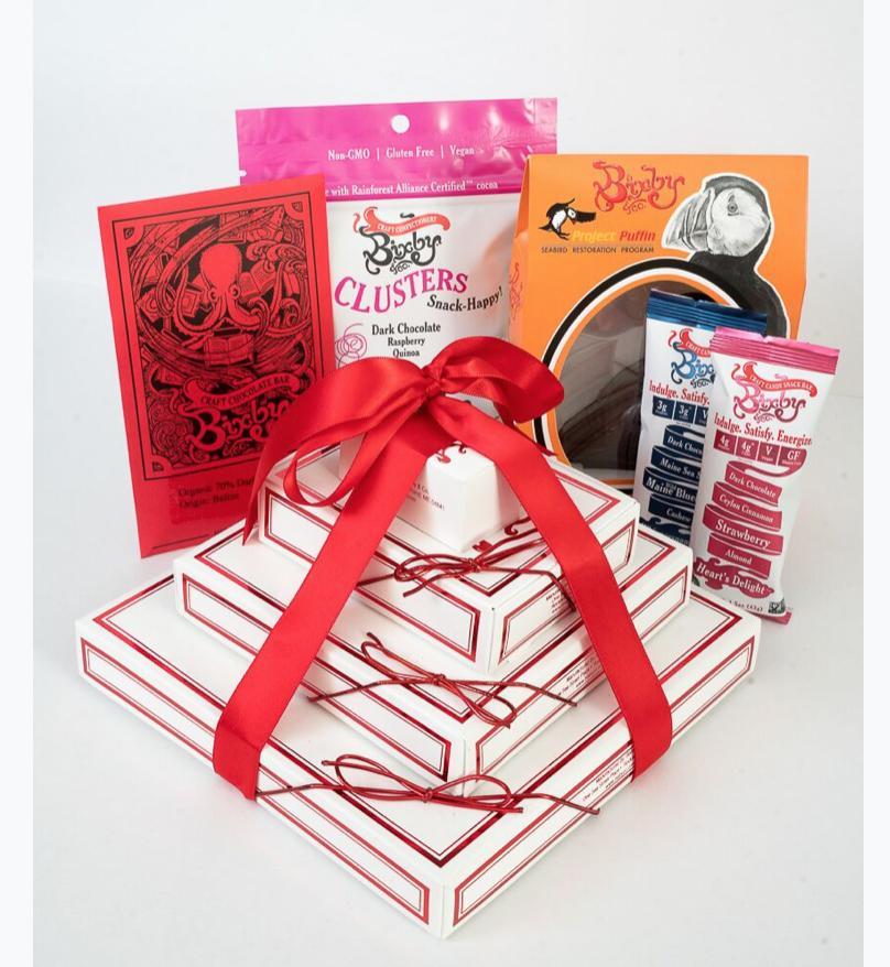 A gift set with stacked white boxes tied with a red ribbon, surrounded by assorted Bixby & Co. chocolate, snack, and bar packages in various colors.