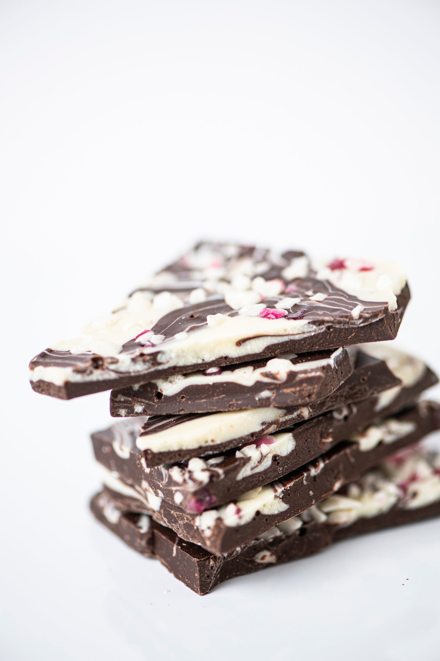 Stack of dark chocolate bark pieces with white chocolate drizzle and bits of red candy on a white background.