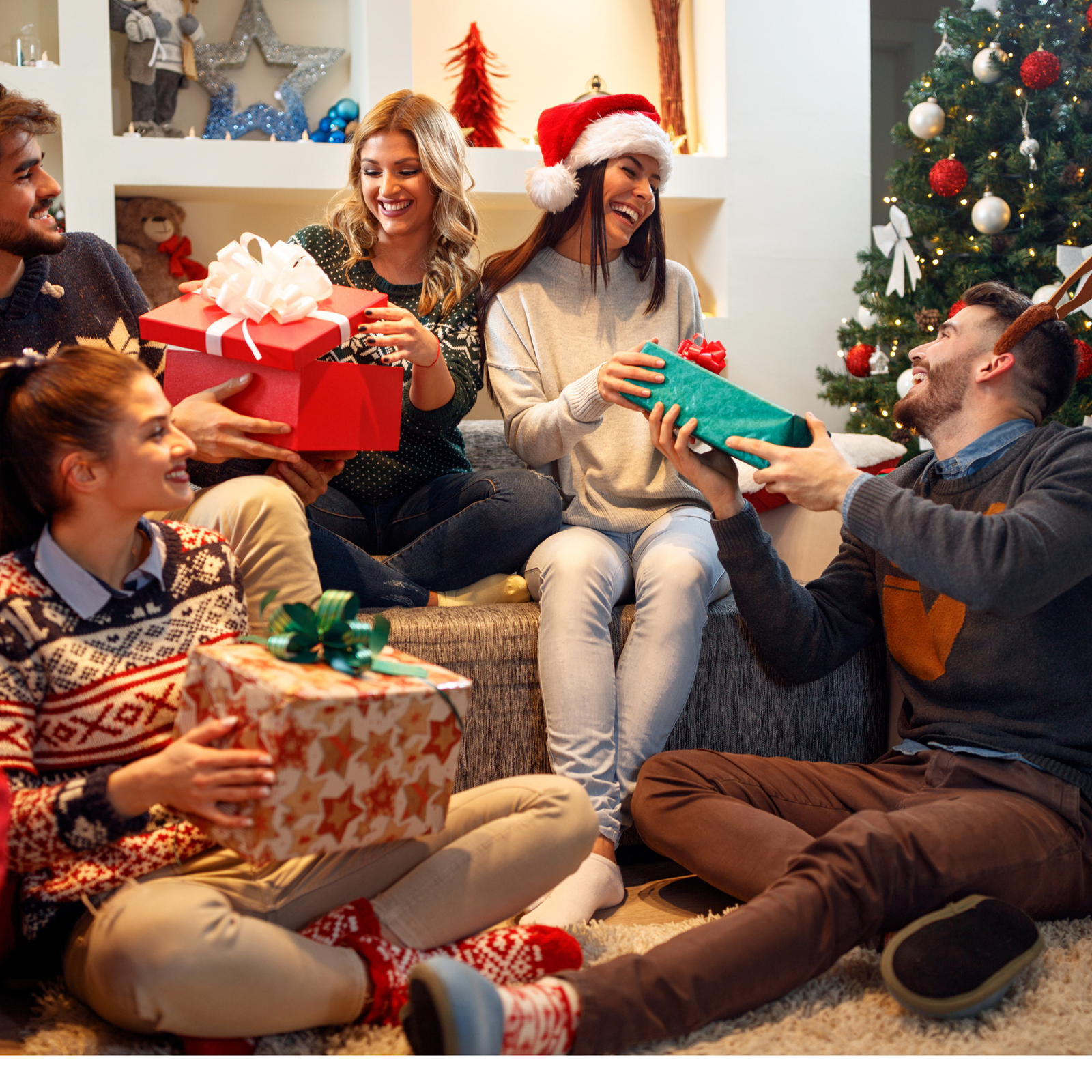 Five friends exchange wrapped gifts and smile together by a decorated Christmas tree and fireplace. One person wears a Santa hat; another has reindeer antlers. The room is festive and cozy.