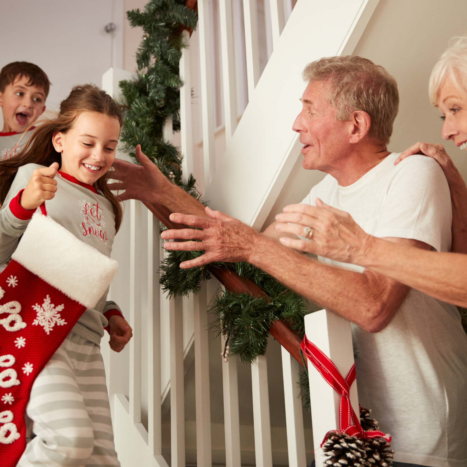 A young girl in pajamas excitedly runs down stairs decorated with garland, holding a Christmas stocking, while two older adults welcome her and a boy follows behind, all smiling joyfully.
