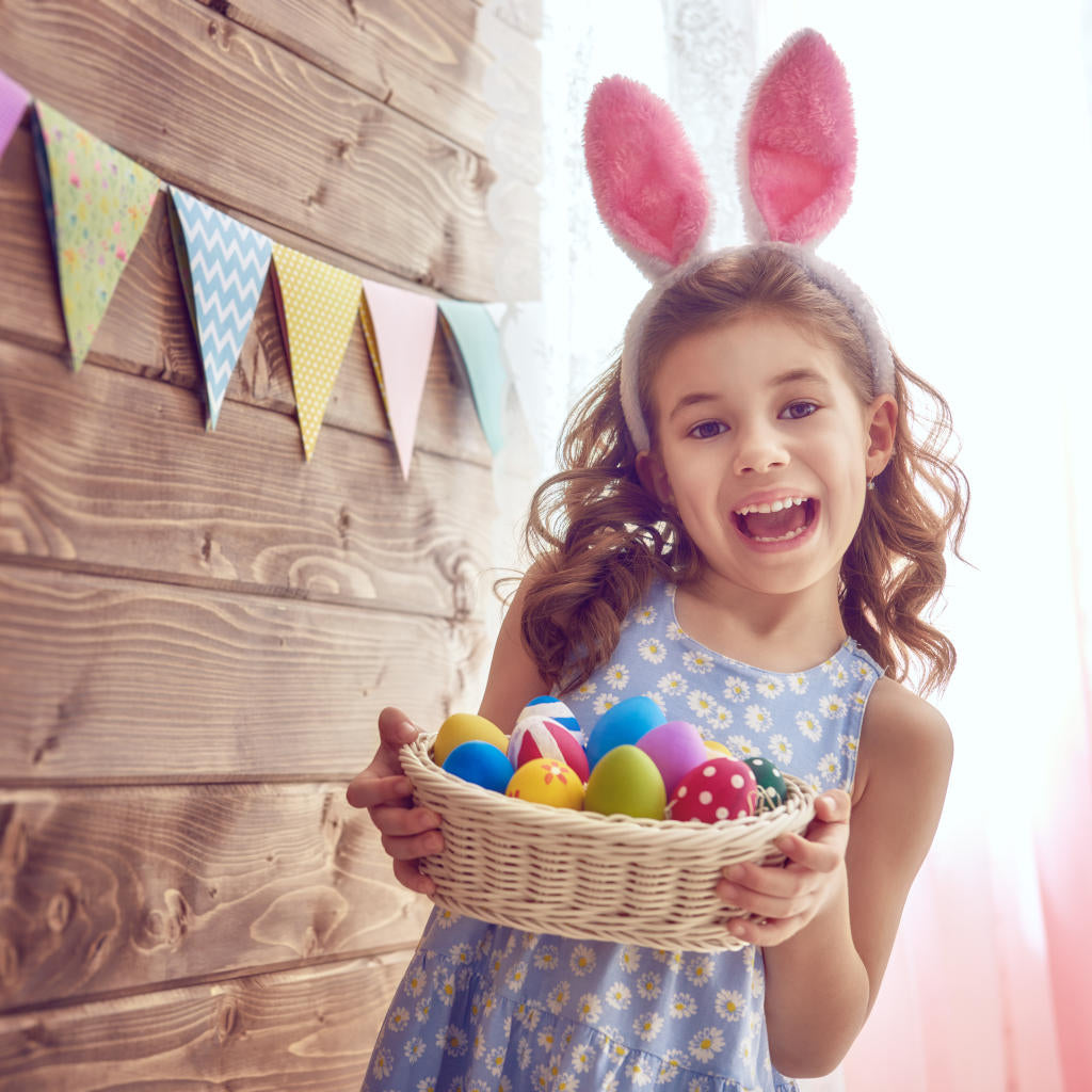 Smiling girl wearing pink bunny ears holds a basket of colorful Easter eggs, standing indoors with pastel bunting and wooden wall in the background.