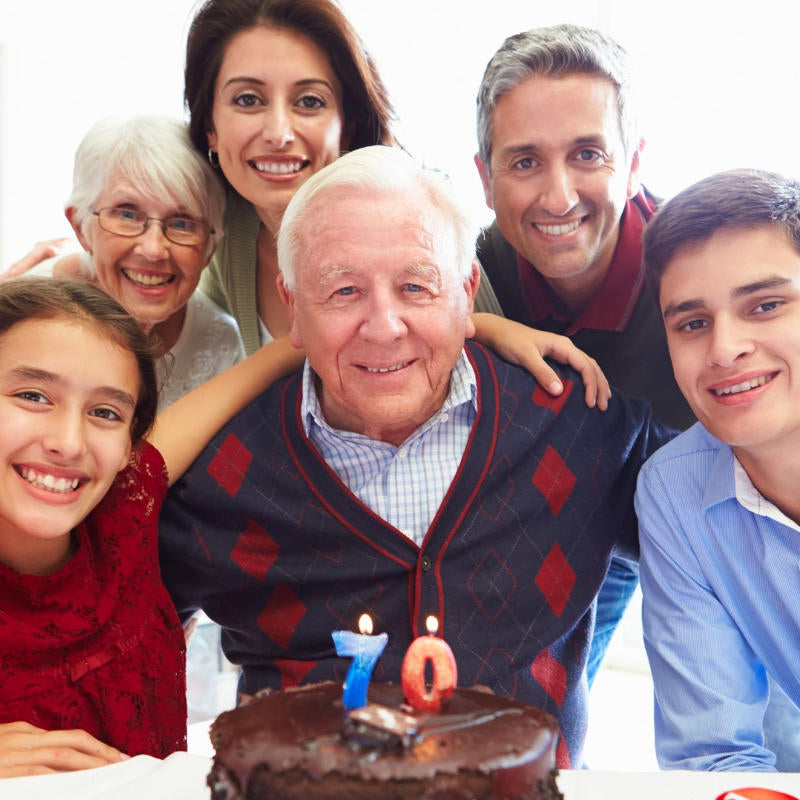 Smiling multigenerational family gathered around an elderly man celebrating his 70th birthday with a chocolate cake and numbered candles.