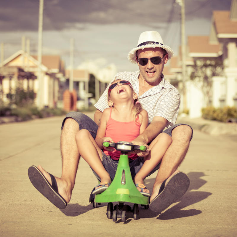 A man and a young girl, both wearing sunglasses and summer clothes, laugh joyfully while riding a small green toy car together on a sunny street in a suburban neighborhood.