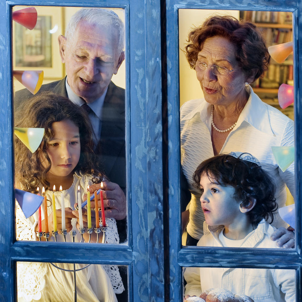 Two children and two elderly adults stand by a window, lighting a menorah for Hanukkah. Colorful decorations hang around them, and the scene is warm and festive inside.