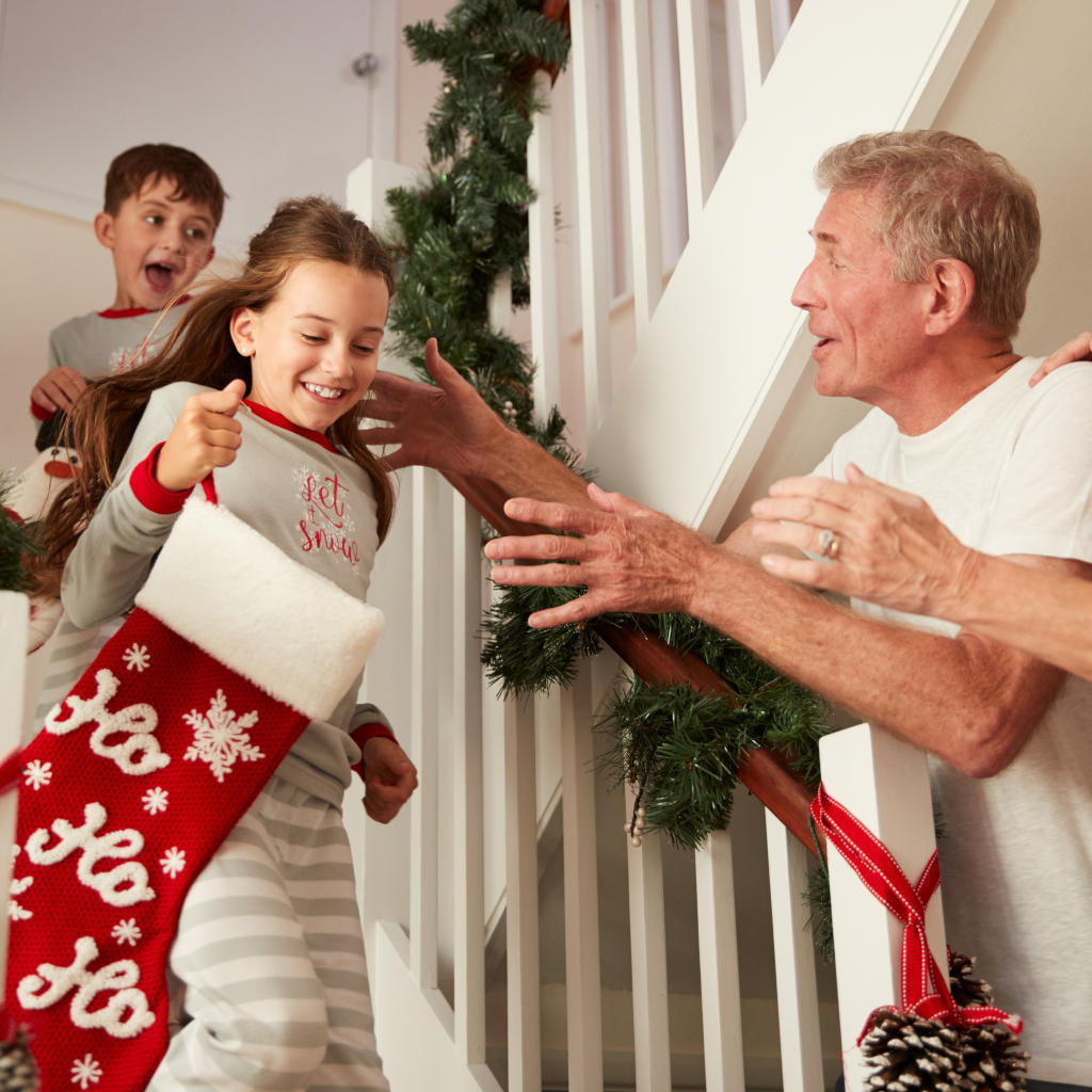 A smiling girl in pajamas runs down the stairs with a Christmas stocking, followed by a boy, as an older man greets them. The staircase is decorated with green garland.