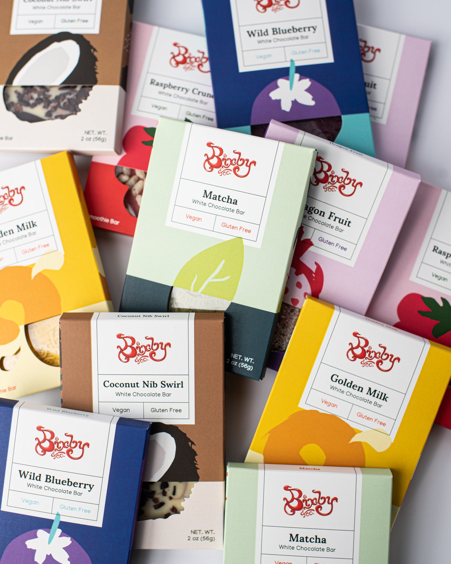An assortment of colorful Bixby & Co. chocolate bar packages, including flavors like Matcha, Coconut Nib Swirl, Raspberry Crunch, and Wild Blueberry, arranged on a white surface.