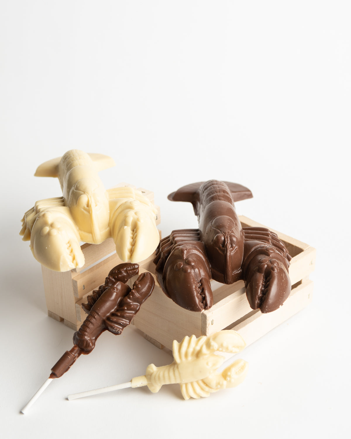 Assorted milk and white chocolate shaped like lobsters and a shark are displayed on small wooden crates, with two lobster-shaped chocolate lollipops on sticks in the foreground.