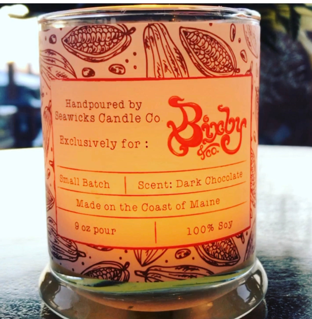 A clear candle jar with cocoa bean illustrations and a label that reads: Handpoured by Seawicks Candle Co. Exclusively for Bixby & Co. Scent: Dark Chocolate, 9 oz pour, 100% soy, Made on the Coast of Maine.