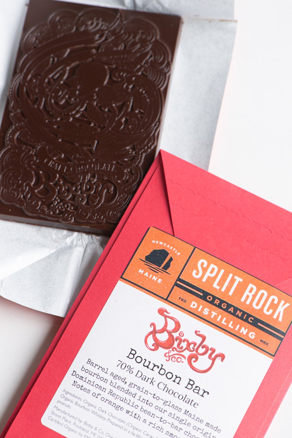 A dark chocolate bar partially unwrapped from foil sits beside a red and white label reading Split Rock Distilling, Bourbon Bar, 70% dark chocolate, Ragged Coast Chocolates.