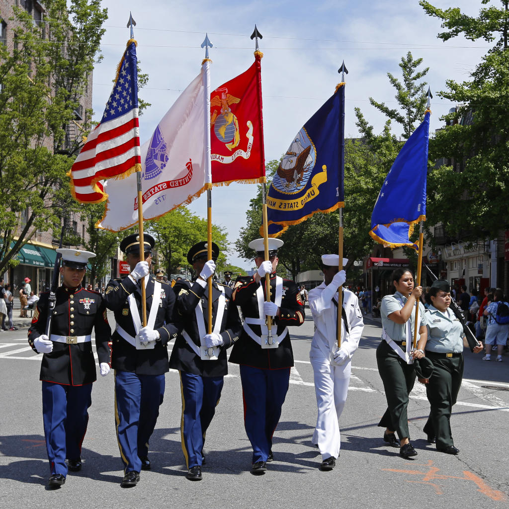 A group of uniformed military personnel carry U.S. and service branch flags while marching in formation down a city street during a parade on a sunny day.