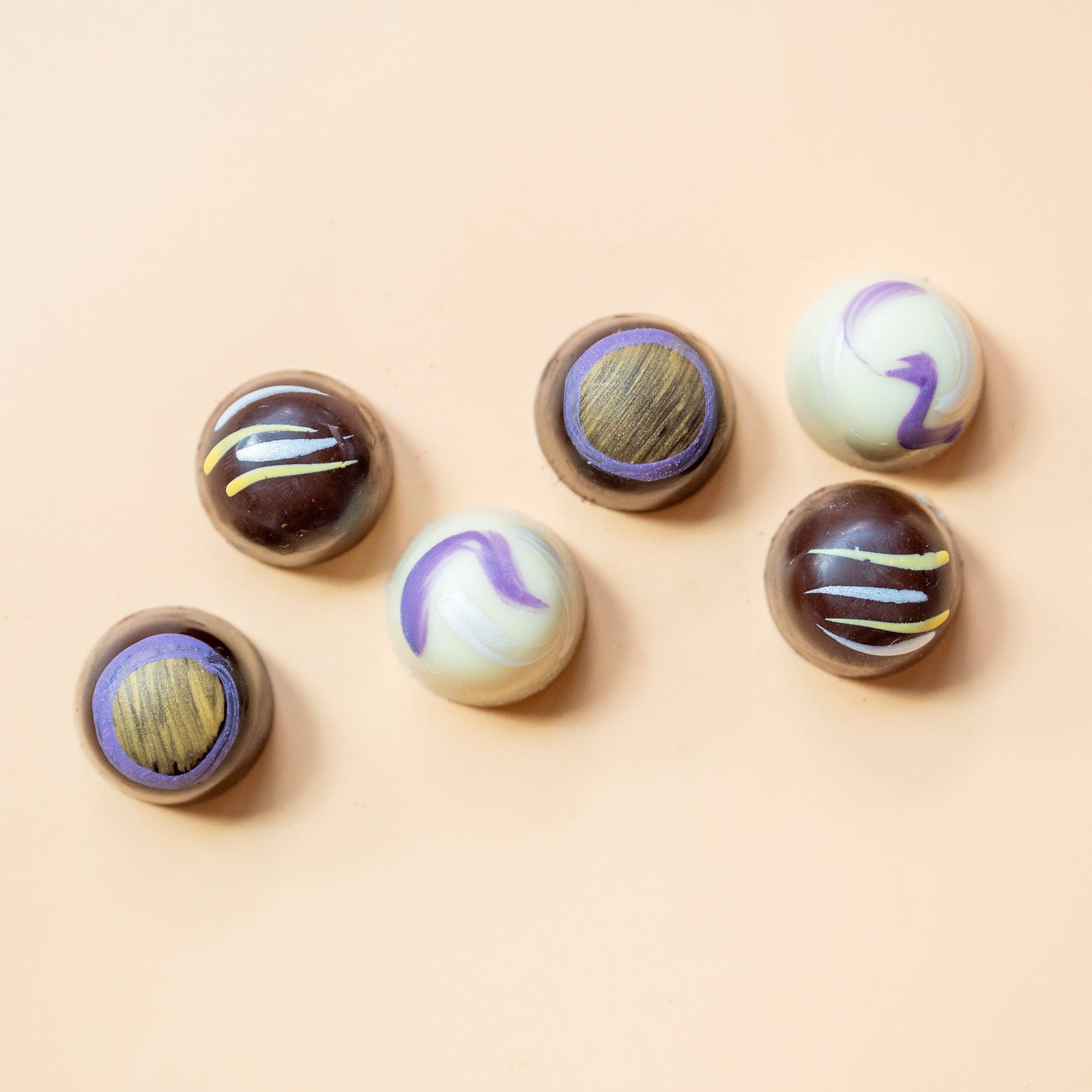 Six artisan chocolates with decorative tops—three with wood-like centers and three with pastel swirls or stripes—are arranged on a light beige surface.
