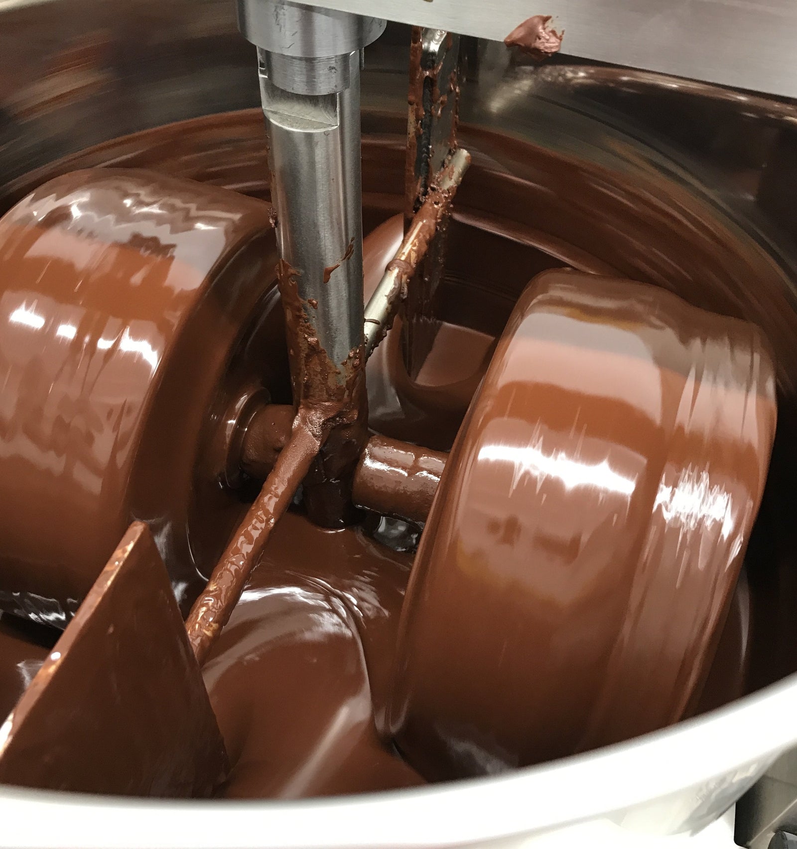 A close-up of a chocolate melanger machine in use, with thick chocolate being ground and mixed between two large rotating stone rollers and a central metal shaft.