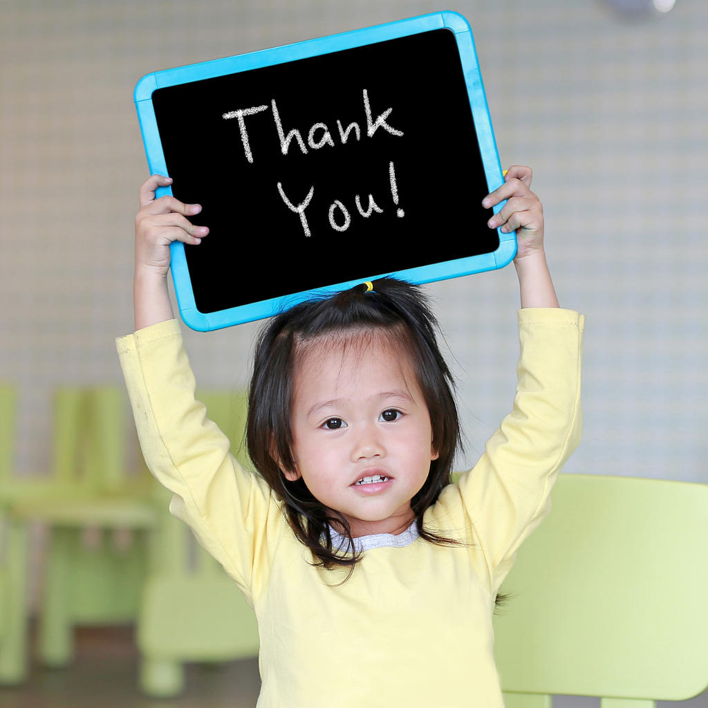 A young child in a yellow shirt holds up a small chalkboard with Thank You! written on it, smiling gently in a brightly lit room with green chairs in the background.