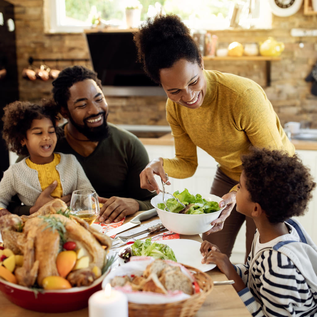 A smiling family of four gathers around a table with a roast turkey and salads, as the mother serves salad to the youngest child in a cozy, sunlit kitchen.