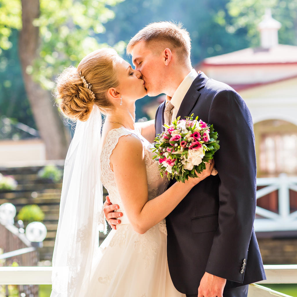 A bride and groom share a kiss outdoors on their wedding day, with the bride holding a bouquet of flowers and sunlight streaming through the trees in the background.