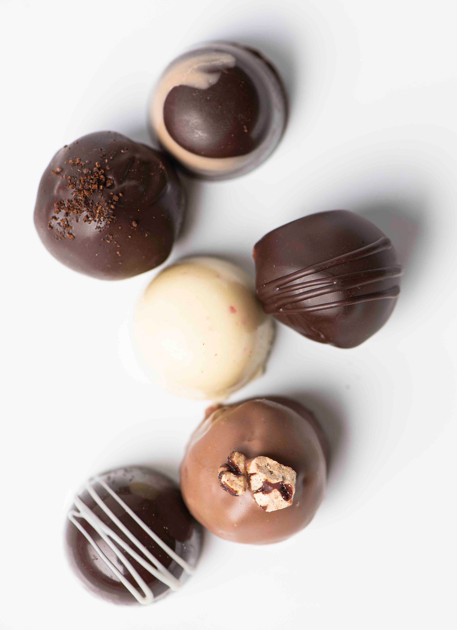 Six assorted chocolate truffles, including dark, milk, and white chocolate varieties, arranged in a loose circle on a white background. Some truffles are decorated with drizzles or toppings.
