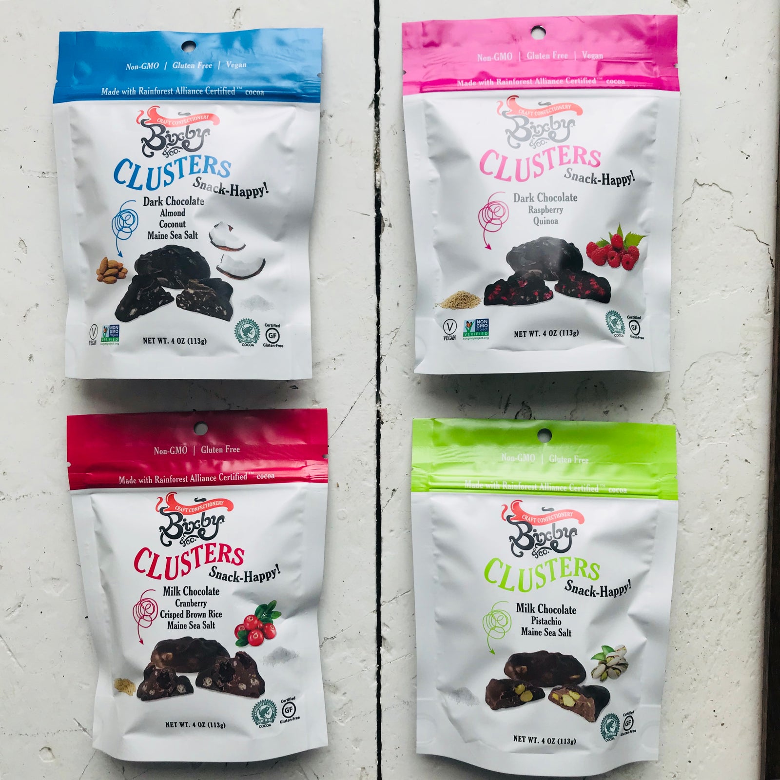 Four bags of bobbySue’s Nuts Clusters snacks in different flavors are arranged on a white background. Each bag has colorful labels: blue, pink, red, and green.