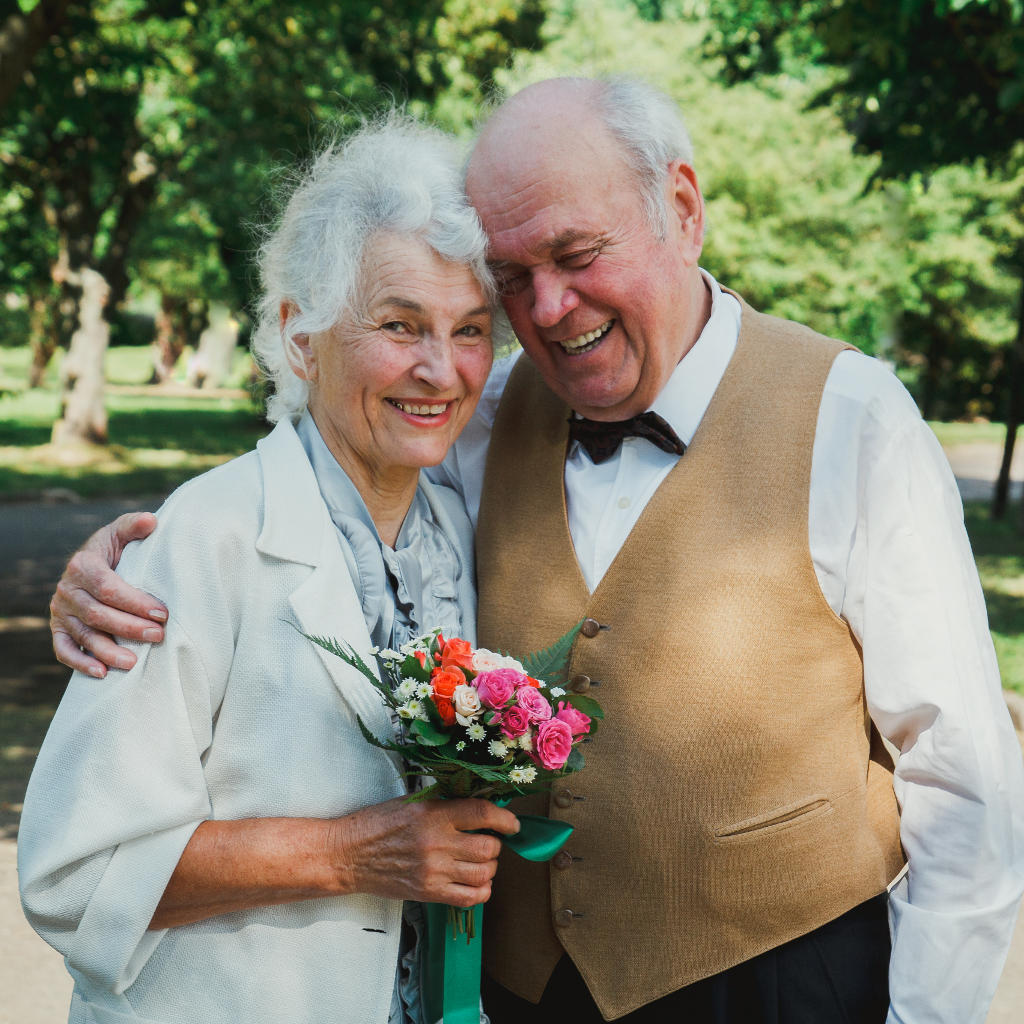 An elderly couple stands close together outdoors, smiling warmly. The woman holds a bouquet of colorful flowers, and the man wears a tan vest and bow tie, both looking happy and affectionate.