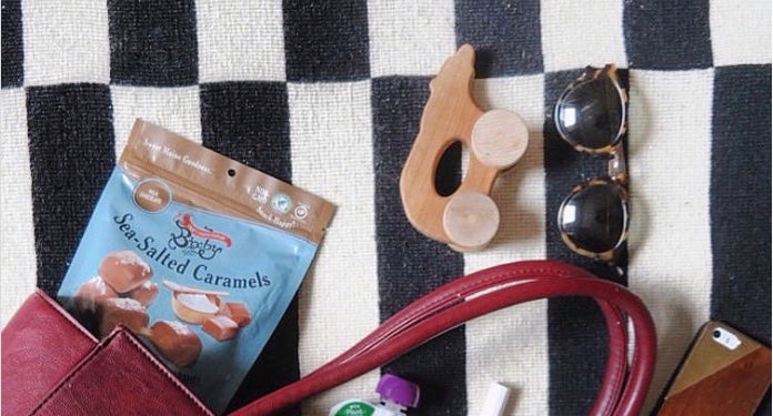 A red purse sits on a black and white checkered surface with sea-salted caramels, wooden toy, tortoiseshell sunglasses, lip balm, and a phone partially visible.