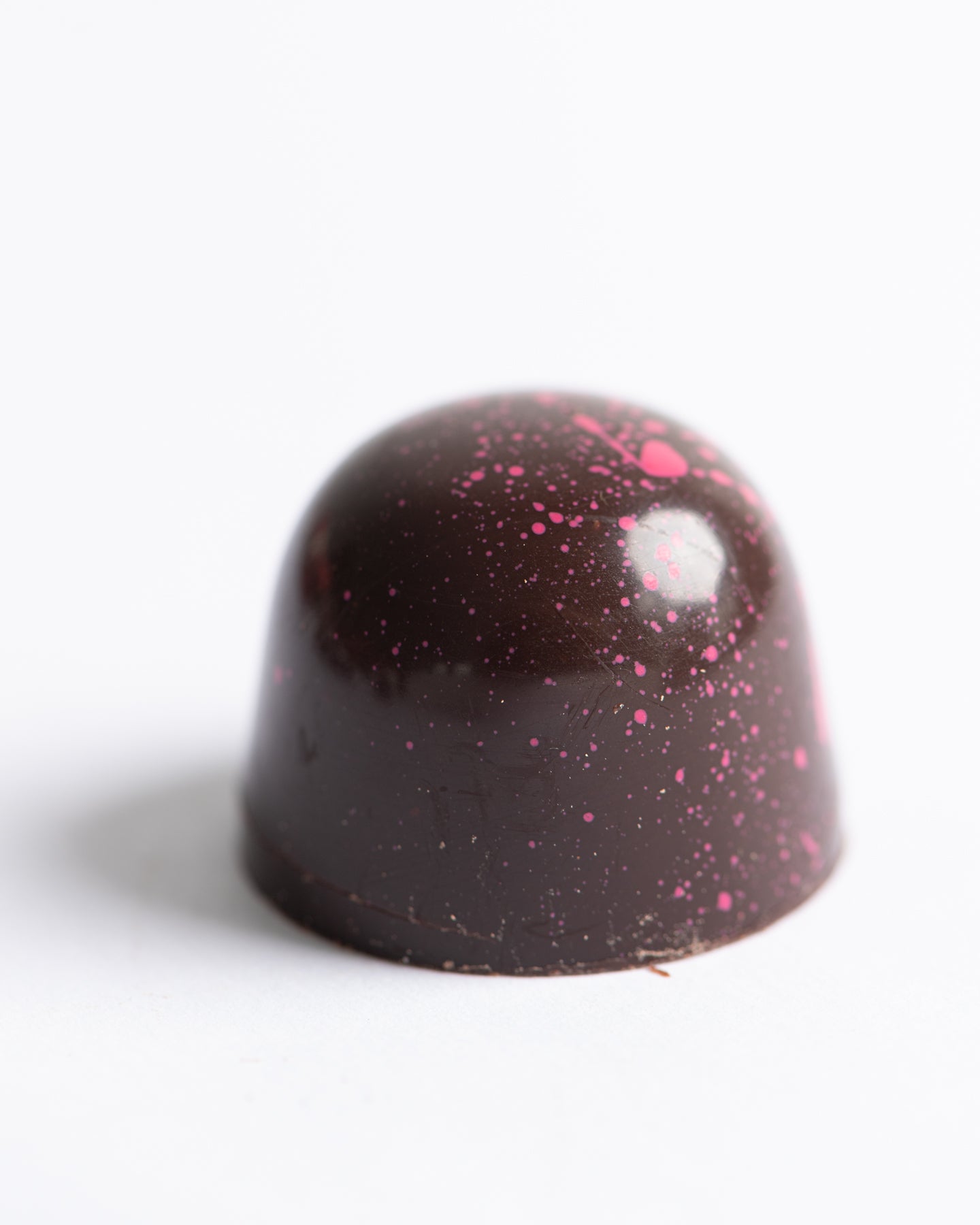 A dome-shaped dark chocolate bonbon with pink speckles sits on a white surface.