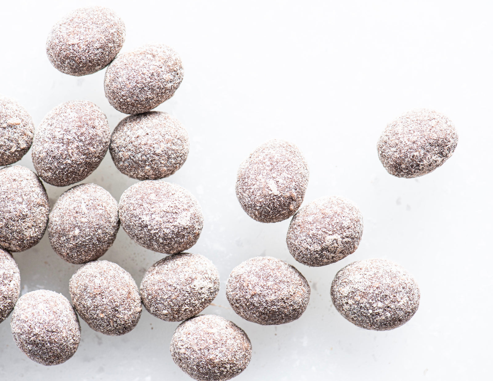 A group of chocolate-coated almonds covered in a light dusting of powdered sugar, scattered on a white surface.