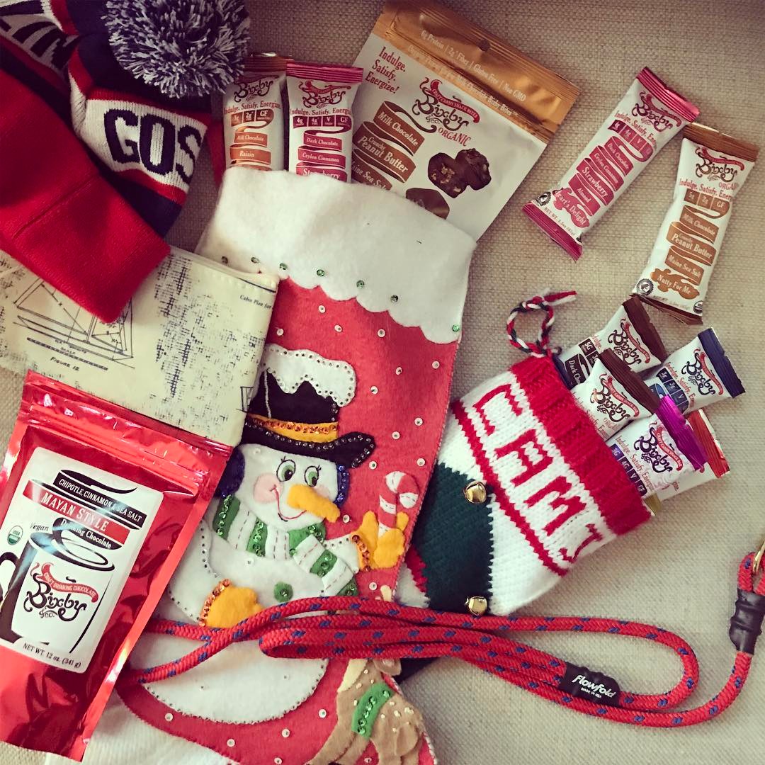 A festive display featuring a snowman stocking filled with snacks, a red knit winter hat, a small “CAM” stocking, dog treats, snack bars, a leash, and an envelope on a light surface.