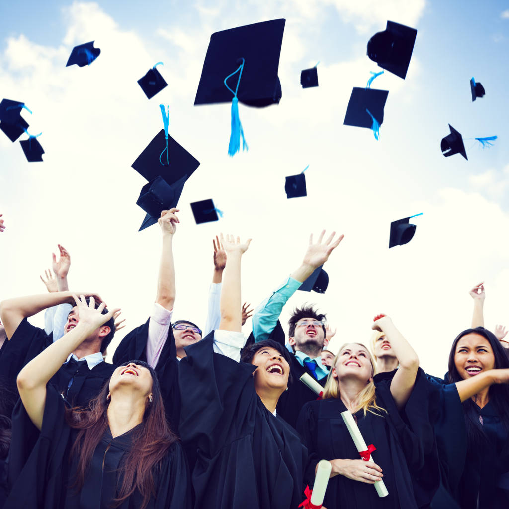 A group of graduates in caps and gowns joyfully throw their graduation caps into the air, celebrating outdoors under a blue sky, holding diplomas and smiling.