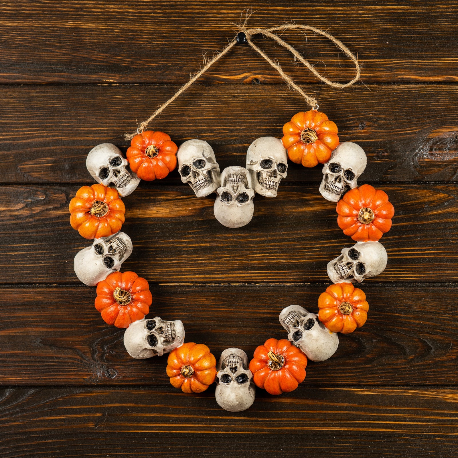 A heart-shaped wreath made of small skulls and orange pumpkins hangs on a wooden background, creating a festive Halloween decoration.