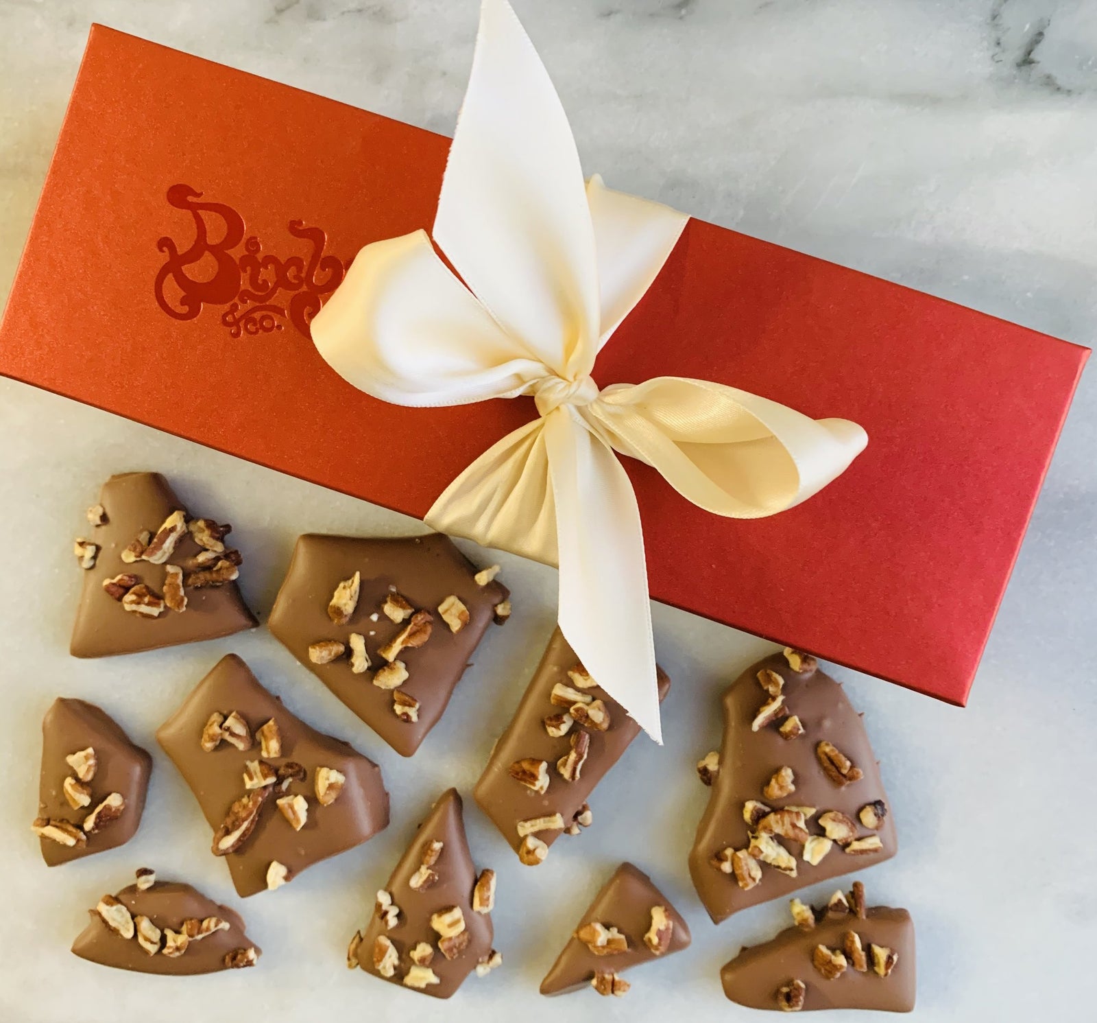 A red gift box with a cream ribbon sits on a marble surface, next to pieces of chocolate toffee topped with chopped nuts.