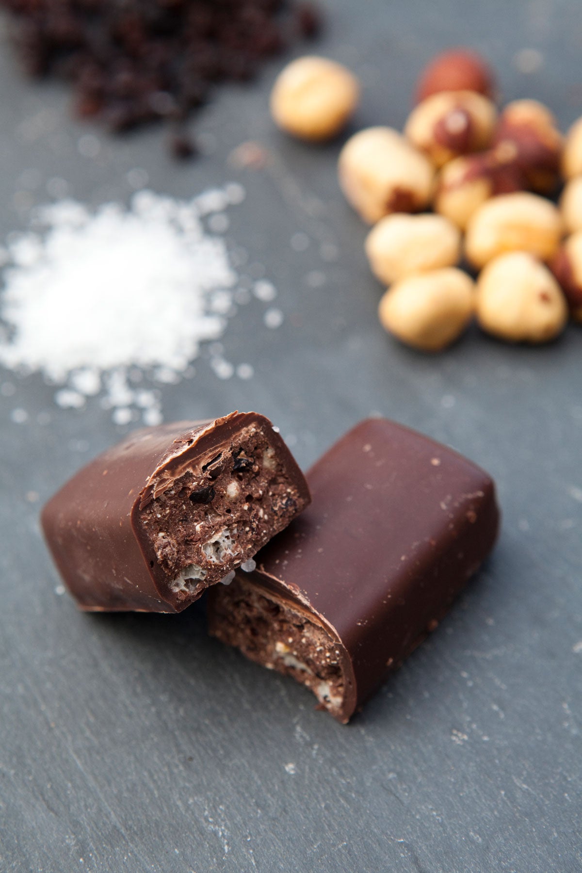 Two pieces of chocolate bars, one cut open to show the inside, are placed on a slate surface with scattered hazelnuts, coarse salt, and cocoa nibs in the background.
