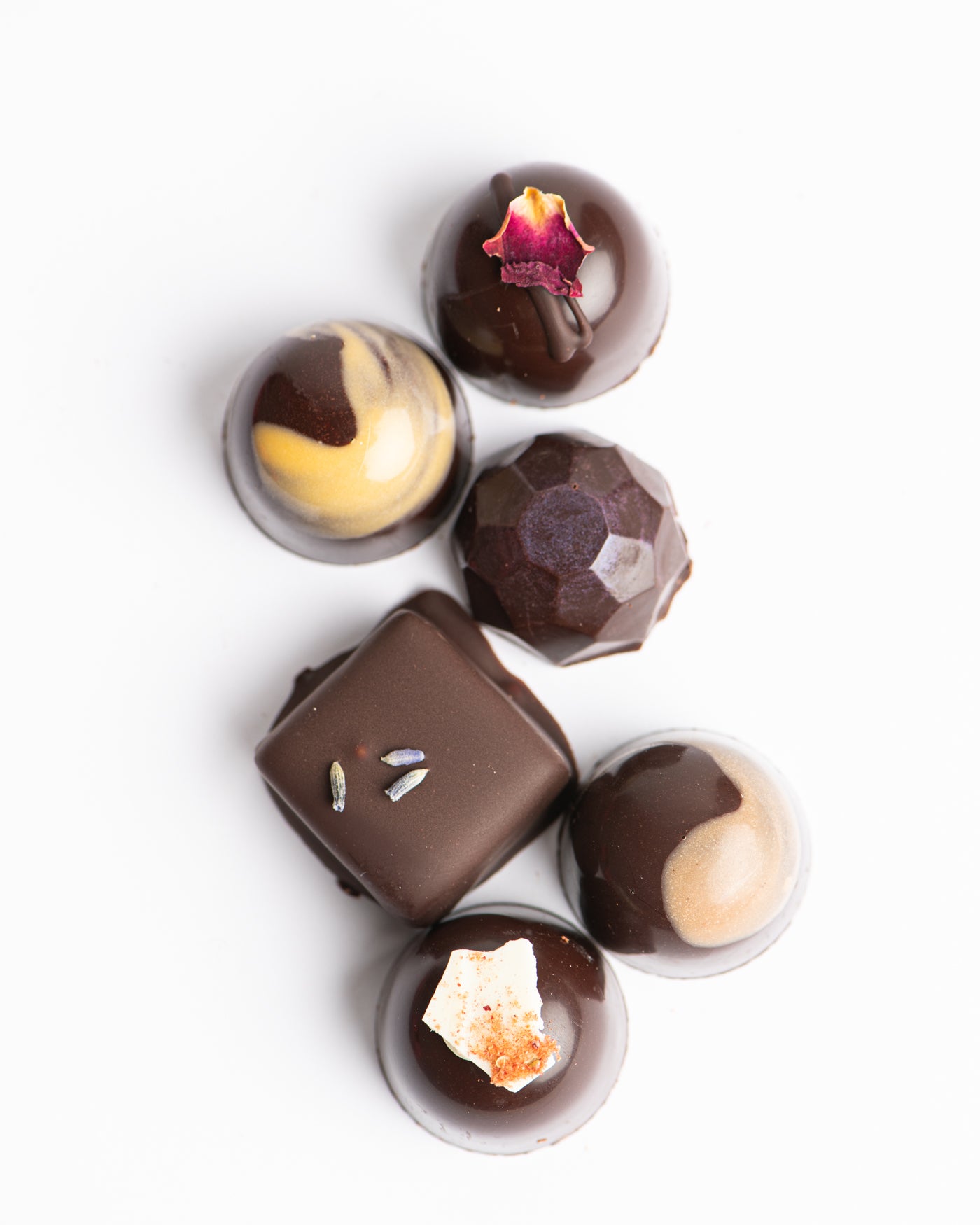 An assortment of six gourmet dark chocolates with decorative toppings and patterns, arranged on a white background. Some have flower petals or dried fruit pieces on top.