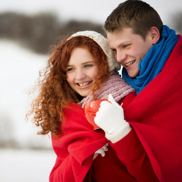 A smiling young couple wrapped in a red blanket stands outdoors on a snowy day. The woman holds a red mug, and both wear winter clothing, looking happy and cozy together.