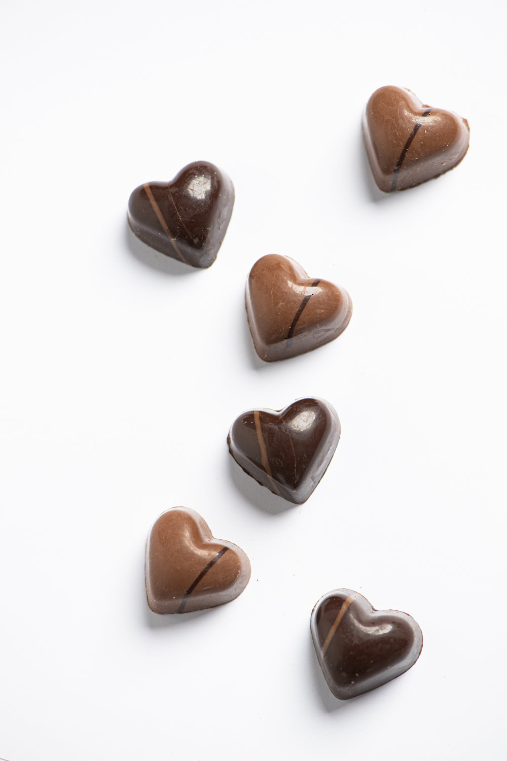 Six heart-shaped chocolates, three dark and three milk chocolate, are arranged on a white background in a loose, diagonal pattern.