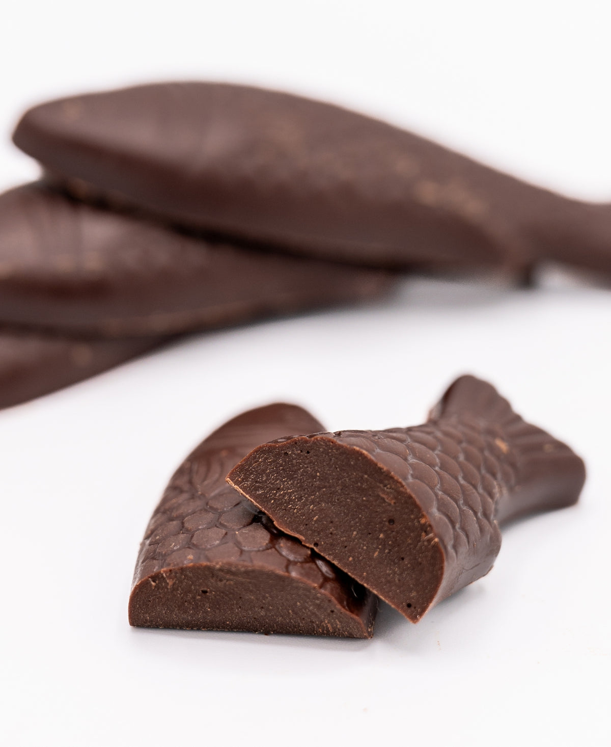 Bixby Chocolates' Chocolate Sardines, Tinned—four artisanal chocolates shaped like fish with detailed scales, one cut to show a smooth interior—all set against a white background. A unique Maine souvenir.