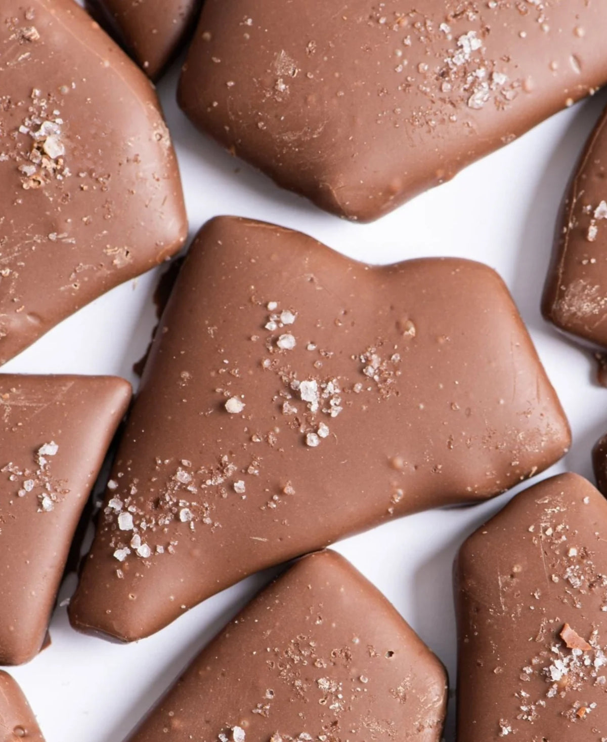 Several pieces of Bixby Chocolates Milk Chocolate Toffee with coarse sea salt are arranged on a white surface. The toffees are irregularly shaped and have a smooth, glossy chocolate coating.