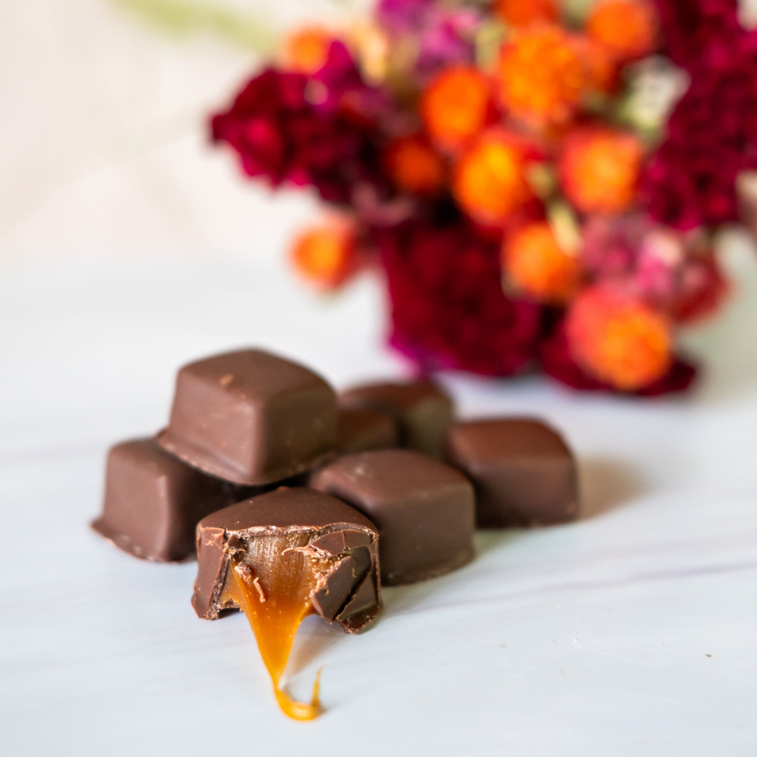A stack of Bixby Chocolates Pumpkin Caramels | 6 Pouches, one with caramel oozing out, sits on a white surface with blurred red and orange flowers in the background—delicious fall treats for the season.