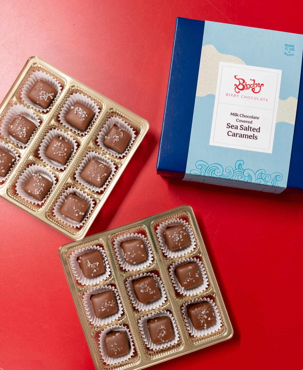 Two open trays of Bixby Chocolates Chocolate Sea Salted Caramels sit on a red surface beside a blue and white box labeled “Chocolate Sea Salted Caramels Gift Box.”.