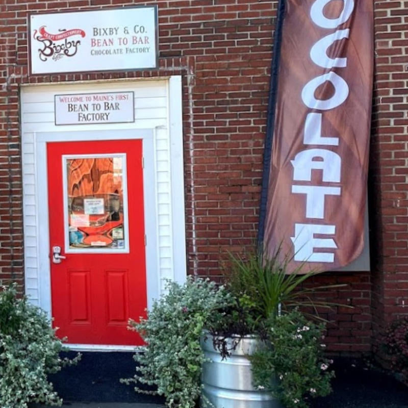 A red door with a sign above reading “Welcome to Maine’s First Bean to Bar Factory” is set in a brick building. A vertical flag says “CHOCOLATE” and potted plants sit near the entrance.