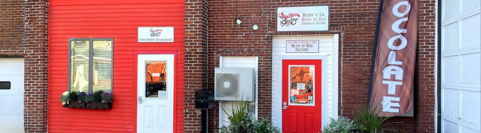A red and brick storefront with two white doors, a large vertical “CHOCOLATE” flag, potted plants by the entrance, and signs for “Bean to Bar Chocolate Factory.”.