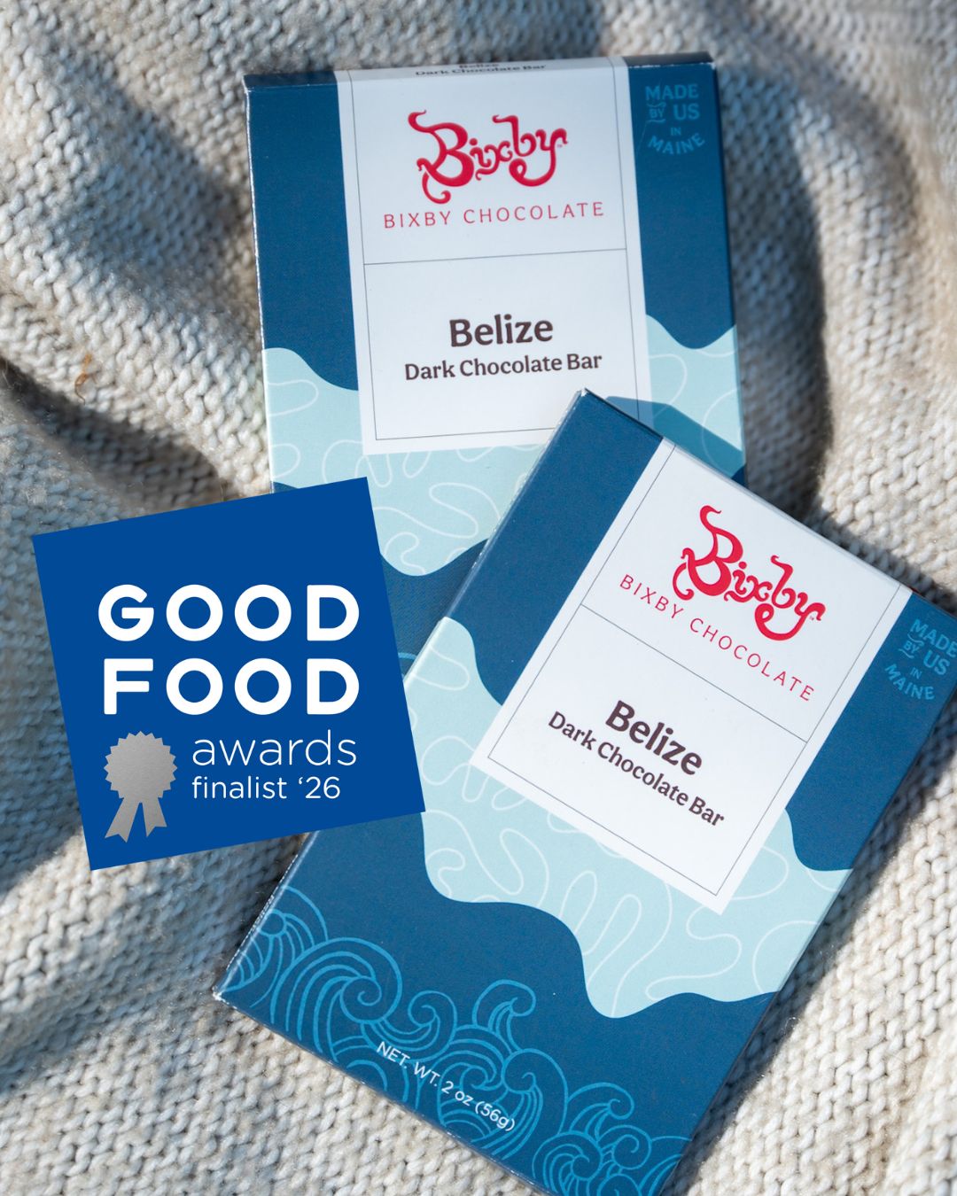 Two blue and white Bixby Chocolates Organic Belize 70% Dark Chocolate Bar boxes are displayed on a beige surface next to a blue Good Food Awards finalist 26 card.