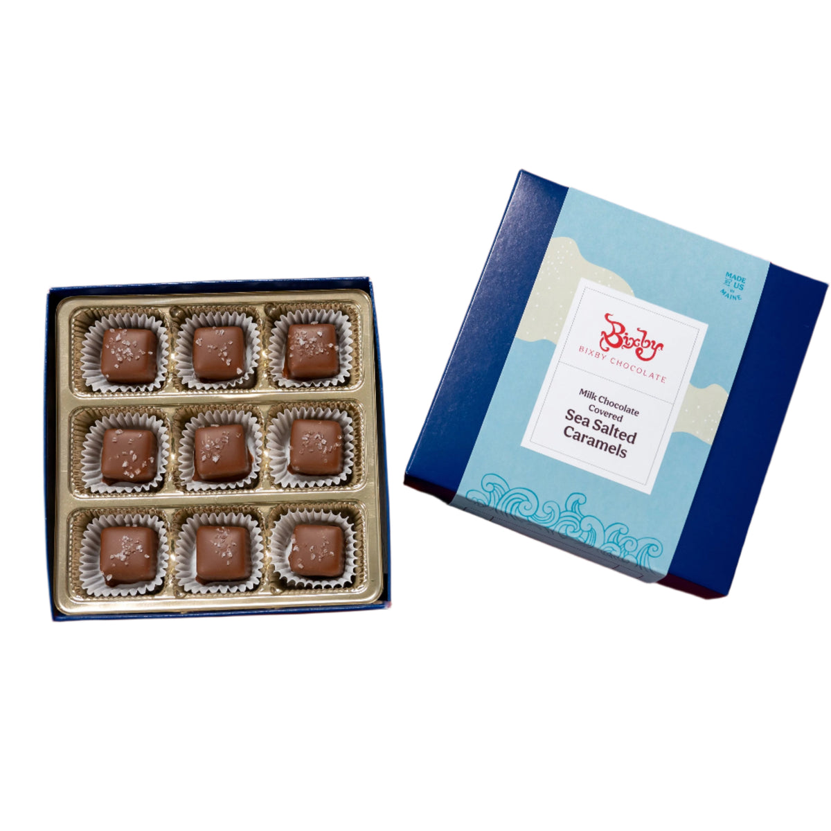 An open box of nine Milk Chocolate Maine Sea Salted Caramels by Bixby Chocolates sits beside its blue and white “Sea Salted Caramel” packaging. Each caramel is wrapped in a paper cup.