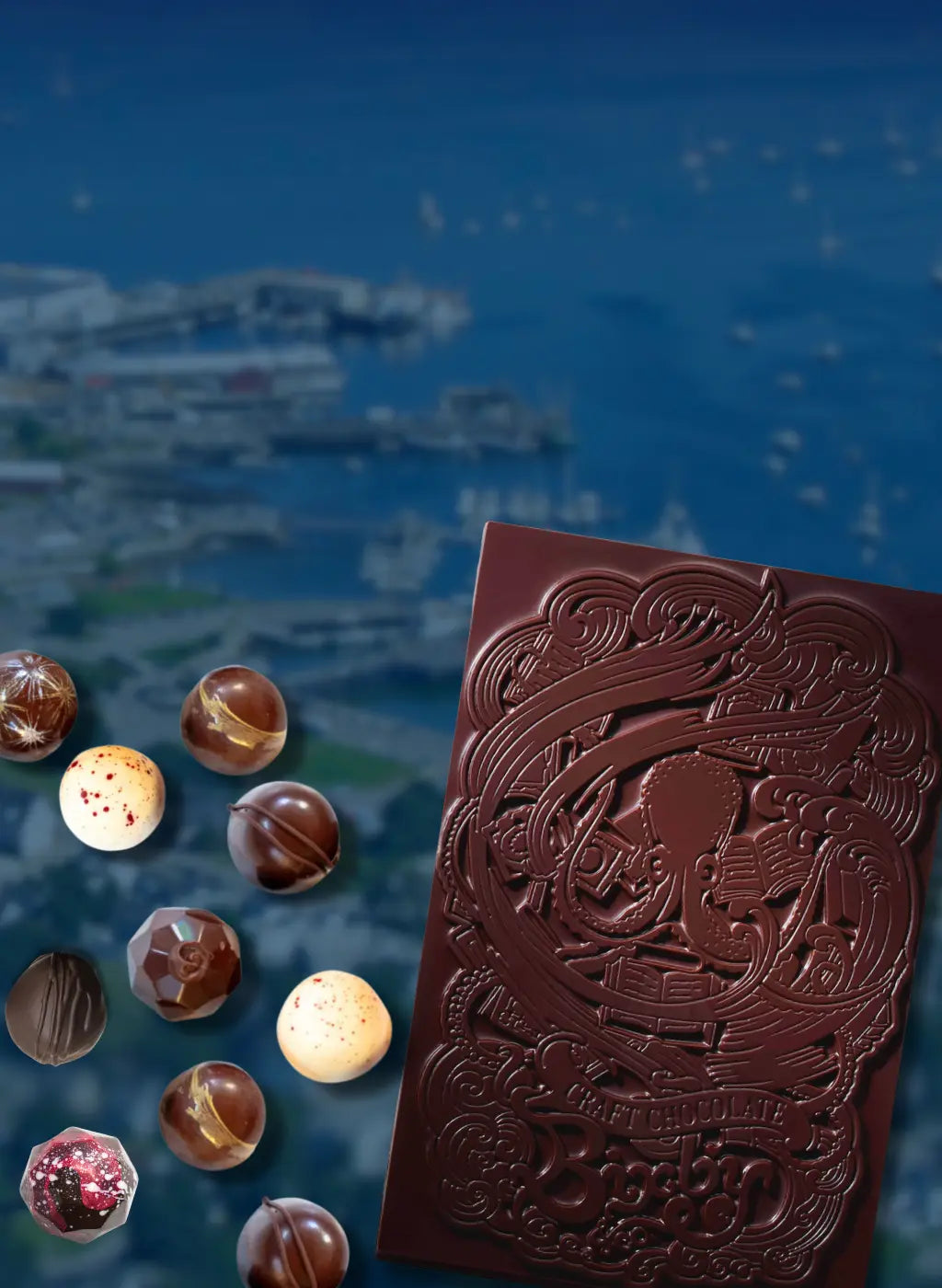 A decorative dark chocolate bar and assorted chocolate truffles are displayed in the foreground, with an aerial view of a coastal city and harbor blurred in the background.