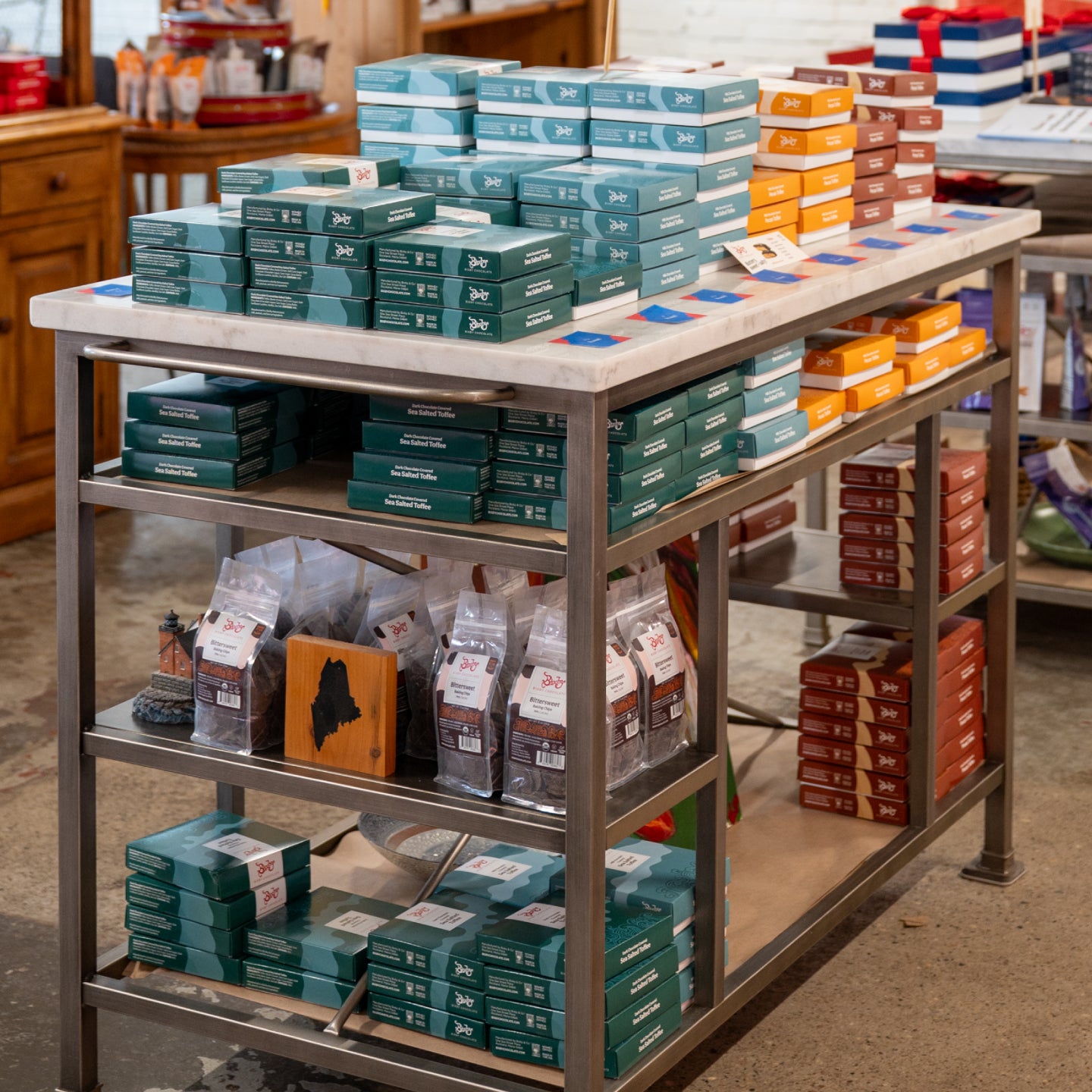 A metal display table in a store holds neatly stacked boxes in teal, orange, and maroon, with several bags of coffee or similar products on the middle shelf and more boxes on the bottom shelf.