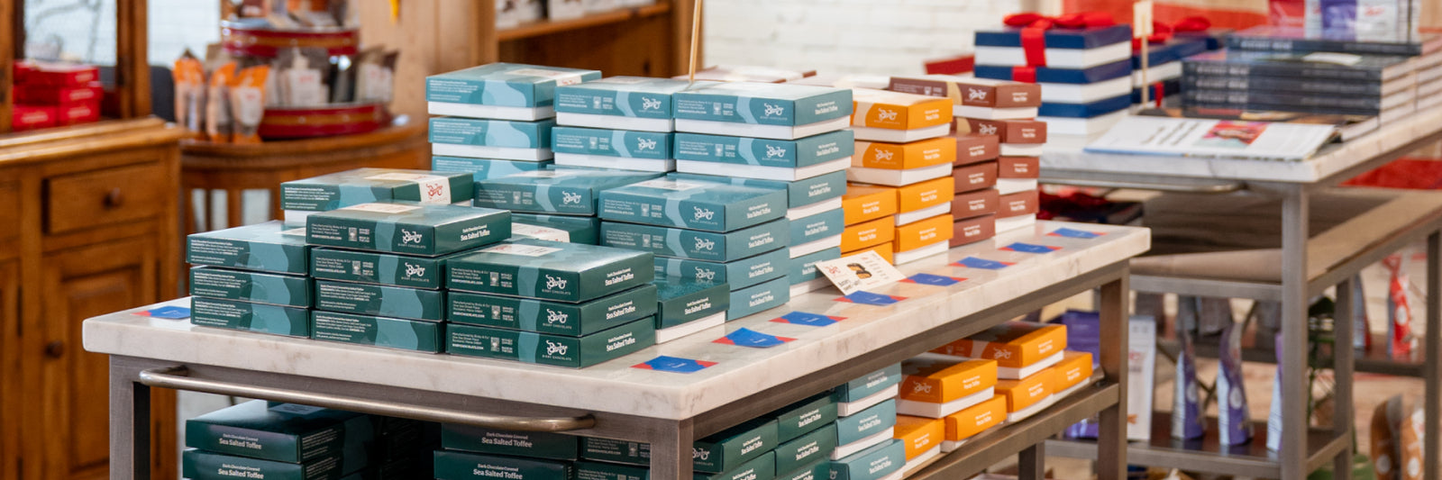 Stacks of colorful rectangular boxes, including teal, orange, and brown, are neatly displayed on a marble countertop in a well-lit store with wooden shelves and additional items in the background.