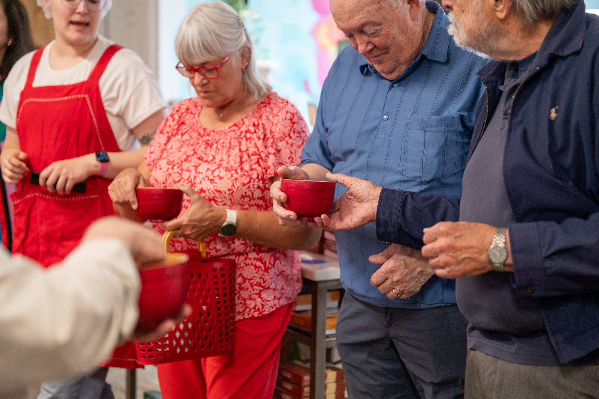 Several older adults stand together indoors, each holding a red bowl. One person wears a red apron, another has a red and white floral top, and they appear to be participating in a group activity or meal.