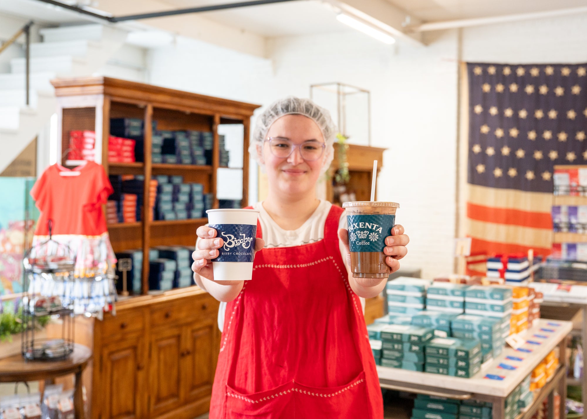 A person wearing a red apron and hairnet holds out two drinks, one in each hand, smiling inside a shop with shelves of colorful boxes and a large American flag on the wall behind them.