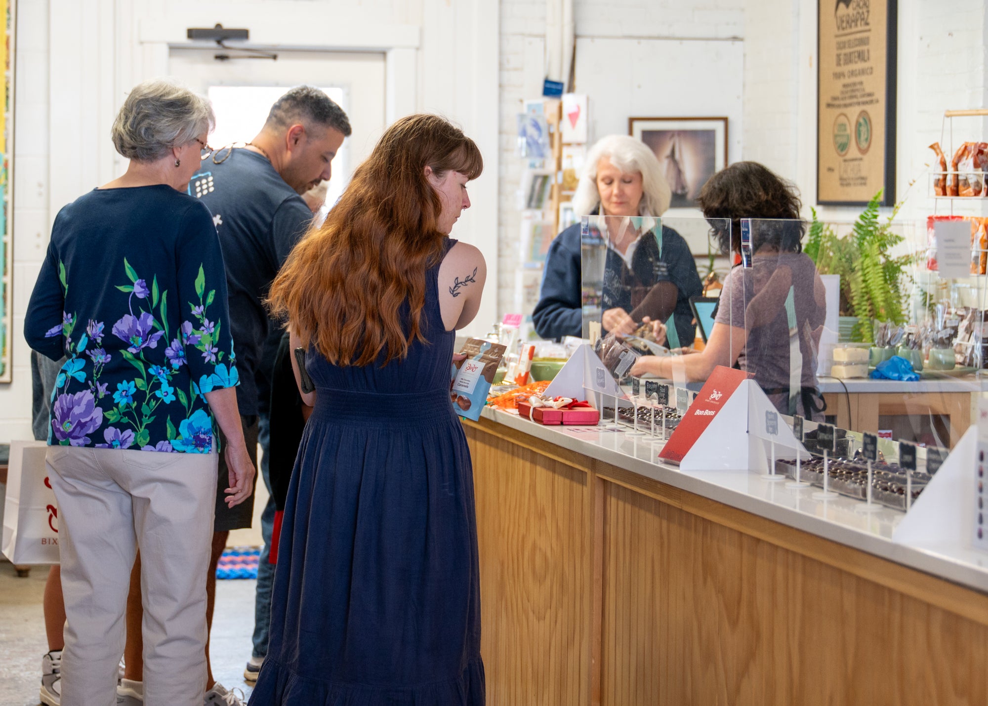 Several people stand in line at a counter inside a shop, waiting to be served by two employees behind a clear protective barrier. One woman holds a drink, and there are various items displayed on the counter.